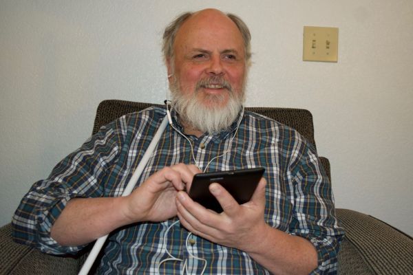 A man sitting in an chair, using the NFB NewsLine application on a tablet.