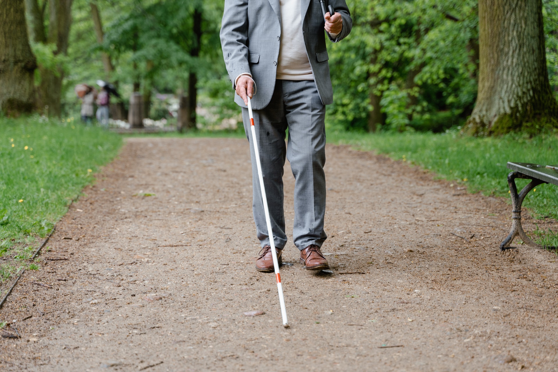 A blind man with white mobility cane, walking down a paved trail in a park.